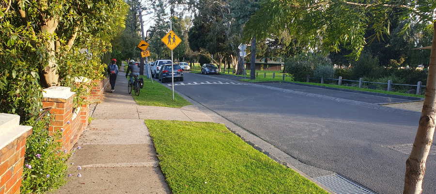 Photo of street in Alphington, showing a footpath with pedestrians, grass verge and road, with cars.